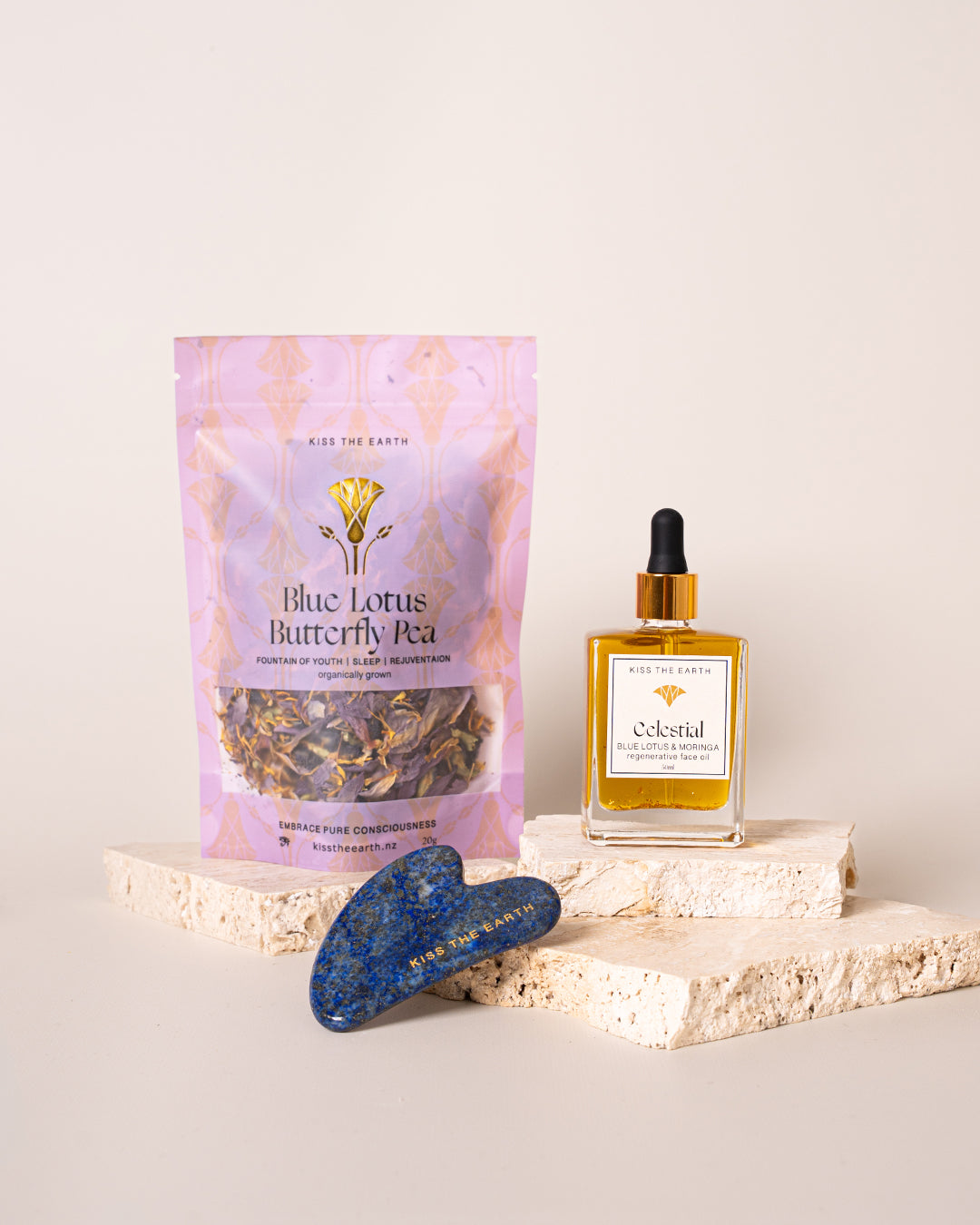 A collection of skincare products including a face oil dropper bottle, a Lapis Gua Sha tool, and a packaged Blue Lotus Butterfly Pea Tea bag, all displayed on a light background.
