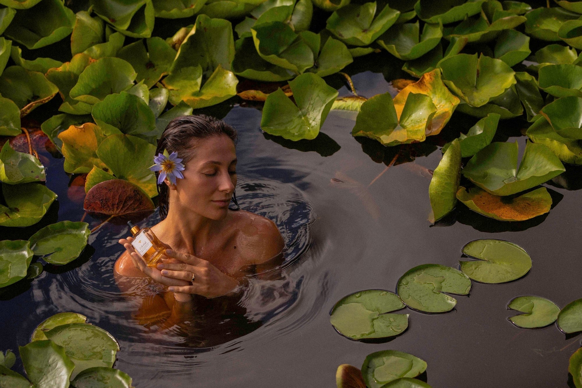 Goddess in a pond holding a blue lotus flower in her head and a bottle of blue lotus natural face oil 